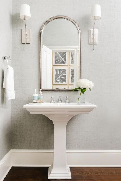 Between nickel sconces mounted to a wall covered in gray wallpaper, an arched beaded mirror hangs over a white pedestal sink finished with a polished nickel cross handle faucet. Powder Room Ideas With Wood Floors, Modern Bathroom Pedestal Sink, Small Powder Bathroom Ideas Pedestal Sink, Mirror For Pedestal Sink, Pedestal Sink Mirror, Mirror Over Pedestal Sink, Staged Bathroom, Powder Room With Pedestal Sink, Pedastal Sink