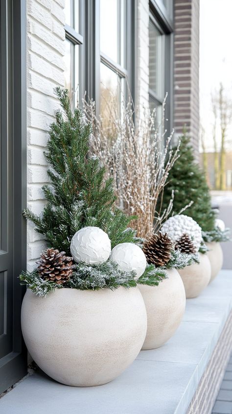 Five large beige planters with pine cones, evergreen branches, and white Christmas balls on a Dutch house's winter porch. Winter Porch Planters, Outdoor Christmas Planters Winter Porch Front Doors, Winter Greenery Outdoor Pots, Winter Planter Ideas Outdoor, Christmas Porch Planter Ideas, After Christmas Winter Decor Porch, Christmas Outdoor Pots Planters, Holiday Planters Front Porches, Winter Porch Pots Diy