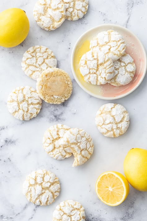 Overhead, scattered Chewy Lemon Almond Crinkle Cookies on a marble surface with a few fresh lemons and three cookies on a small ceramic plate. Lemon Turmeric Crinkle Cookies Nyt, Lemon Powder Cookies, Almond Egg White Cookies, Lemon Amaretti Cookies, Lemon Turmeric Crinkle Cookies, Lemon Tumeric Cookies, Meyer Lemon Cookies, Sweet Egg White Recipes, Recipes With Almond Extract