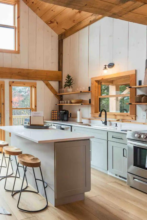 Kitchen of a post and beam cabin with light blue wooden cabinets, white painted wood walls, a white marble top island, stools with wooden seats, and a chrome oven. Cabin Lake House Interior, Woodland Cabin Interior, Modern Cozy Cabin Interior, Bright Cabin Interior, Rustic Beach Cabin, Open Concept Cabin, Post And Beam Cabin, Cabin Home Ideas, Minnesota Cabin