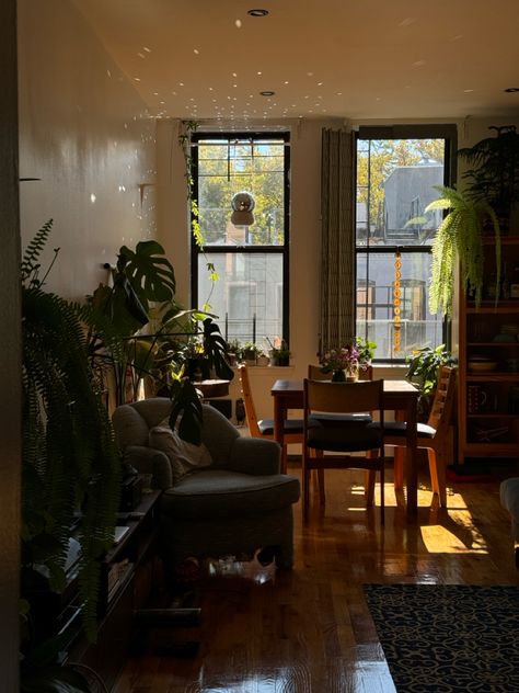 Warm toned south facing living room with sunlight streaming in. Disco ball hangs in window and reflects light across the ceiling & walls. Room is filled with plants, ferns, monstera, cactus & pothos. Small lounge chair separates living space in the foreground from dining table & glass cabinet in. Nyc Apartment Plants, My Dream Life Aesthetic, Dream Life Aesthetic, Apartment Plants, Apartment Vibes, Life Aesthetic, Nyc Apartment, My Dream Life, Cozy Living Rooms