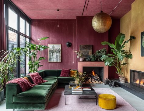 This living room is a bold study in color contrasts and texture. The rich, burgundy walls paired with the deep green velvet sofa create an opulent yet inviting atmosphere. Warmth radiates from the fireplace, while the lush plants and gold accents bring nature and luxury into perfect harmony. Every detail, from the oversized pendant to the pops of mustard and pink, makes this space feel both eclectic and elegant. It’s a true celebration of daring design, where each piece stands out yet fits se... Dark Green Velvet Sofa Living Room Ideas, Deep Green Sofa, Accent Wall For Dark Room, Bright Bold Living Room, Contrast Fireplace, Green Sofa Pink Walls, Color Sofa Living Room, Green Burgundy Living Room, Burgundy And Blush Living Room