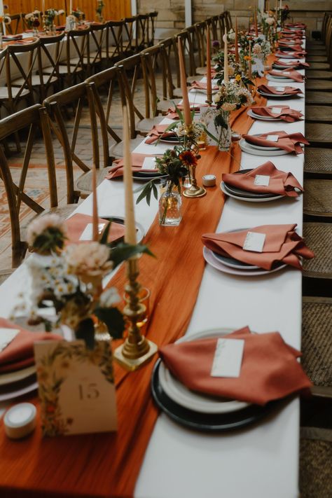 Wedding reception table decor with long burnt orange table runner and burnt orange napkins. Taper candles stand in brass candle holders & elegant Autumn flowers sit in small glass bud vases. Long trestle tables with cross back chairs. White And Burnt Orange Wedding Table, Rustic Wedding Decor Autumn, Burnt Orange Table Runner Wedding, Fall Wedding Table Linens, Wedding Reception Table Runners, Burnt Orange And Brown Wedding Decor, Rustic Wedding Orange, Autumn Wedding Diy Decorations, Fall Wedding Simple Table Decor