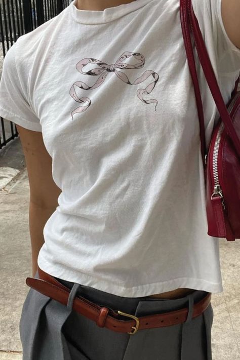 A cropped photo shows a girl from the waist to just below her neck, wearing a white tee shirt with a light pink bow illustration, paired with gray slacks, and a brown leather belt, and cherry red handbag Coquette Graphic Tee, Graphic Baby Tee Outfit, Baby Tees Outfit, Baby Tees Aesthetic, Baby Band Tees, Cute Baby Tees, Baby Tee Aesthetic, Aesthetic Ribbon, Graphic Tee Aesthetic