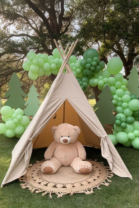 One Happy Camper first birthday party setup featuring a beige teepee with a large teddy bear sitting inside. The backdrop includes green balloon garlands and tree cutouts, creating a perfect outdoor themed photo spot for a camping-themed 1st birthday celebration. One Happy Camper Games, One Happy Camper Centerpieces, One Happy Camper Decorations, Camper Smash Cake, Happy Camper Smash Cake, One Happy Camper First Birthday Girl, Happy Camper First Birthday Boy, One Happy Camper Smash Cake, Wilderness Birthday Party