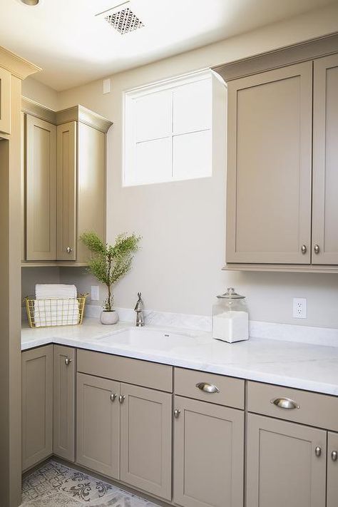 Light brown shaker cabinets accented with satin nickel cup pulls and a white quartz countertop are fitted with a sink matched with a satin nickel faucet located beneath a window flanked by light brown upper cabinets. Brown Painted Cabinets, Light Brown Cabinets, Light Brown Kitchen Cabinets, Light Brown Kitchen, Neutral Kitchen Colors, Taupe Kitchen Cabinets, Beige Kitchen Cabinets, White Kitchen Countertops, Taupe Kitchen