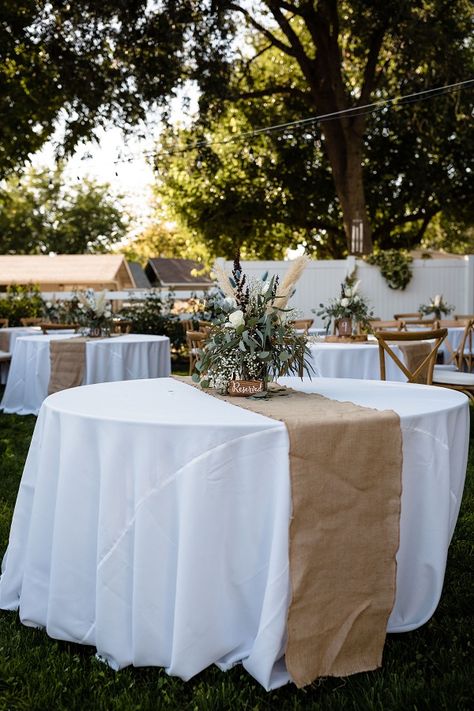 White tablecloths are decorated with beige table runners and floral arrangements in a Chico, CA backyard wedding White Tablecloth With Burlap Runner, Table Runner On White Tablecloth, Wedding Table Covers Ideas Tablecloths, White And Beige Table Decor, White And Wood Party Decor, Beige And White Decor Party, Round Party Tables, White Table Cloth With Burlap Runner, White Table Clothes Wedding