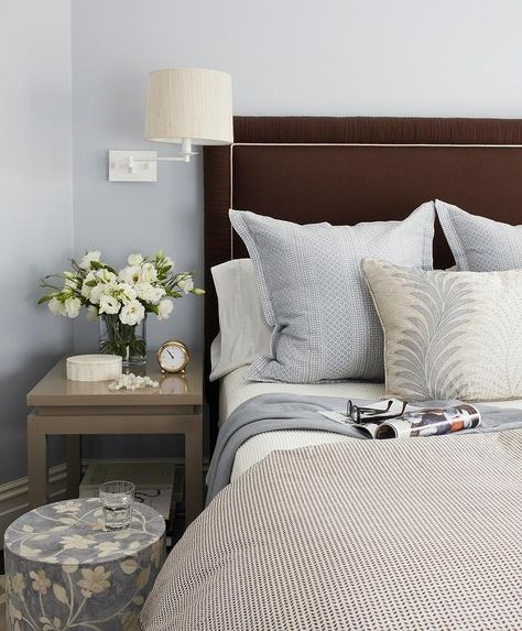 A two tier gray nightstand sits illuminated by a white swing arm sconce mounted on blue walls beside a circular blue floral print stool and a chocolate brown upholstered headboard supporting a bed dressed in a white and black duvet highlighting white shams and blue accent pillows, and a beige, blue and white reef printed lumbar pillow. Brown Headboard, Bedside Table Styling, Blue Headboard, Bedroom Kid, Brown Decor, Black Headboard, Transitional Bedroom, Headboard Wall, Brown Bedroom