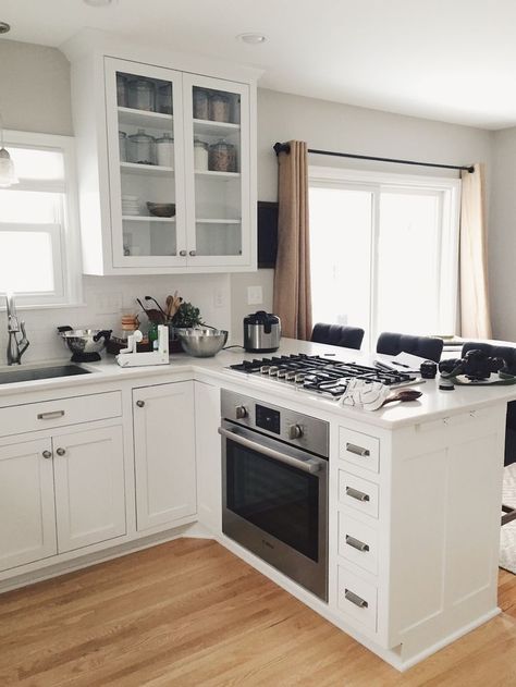 White kitchen with glass door display cabinet and peninsula with oven and cooktop. Kitchen Layout With Peninsula, Mv Ideas, Island With Stove, Kitchen Island With Stove, Craftsman Remodel, Kitchen With White Cabinets, Decor Cabinet, Kitchen Peninsula, Best Kitchen Design