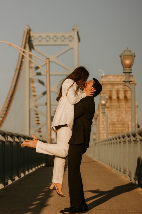 Couple poses for photo on the Ben franklin bridge in Philadelphia to celebrate their engagement. Man in black suit lifts woman in white suit up for a kiss, her foot is popped. Philly Photoshoot, Philadelphia Engagement Photos, Classy Engagement Photos, Engagement Photos Couple, Nyc Engagement, Pictures Engagement, Photoshoot Engagement, Vintage Photoshoot, Engagement Photos Ideas