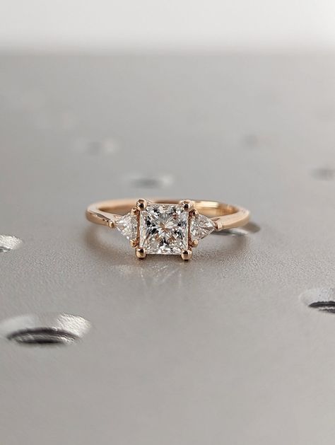 Princess Cut Lab Diamond Ring- 18K Rose Gold- 0.5CT Gemstone Engagement Ring For Women- Dainty Promise Ring- Anniversary Gift- Gift For Her --- Pictures show the 18K Rose Gold with 0.5ct option --- The contemporary basket setting of this elegant ring is supported by an open gallery and framed with trillion diamond or moissanite accents. MAIN STONE Type: Lab Grown Diamond Cut: Princess Color: D-F Clarity: VVS-VS SIDE STONES Type: Natural Diamond / Moissanite Cut: Triangle Number of Gemstones: 2 M Engagement Ring Gold Princess Cut, Princess Cut Engagement Ring With Side Stones, Square Shaped Rings Engagement, Moissanite Engagement Ring Princess Cut, Princess Cut 3 Stone Engagement Ring, Gold Princess Engagement Ring, Unique Square Cut Engagement Rings, Princess Cut Promise Ring, Vintage Square Engagement Rings