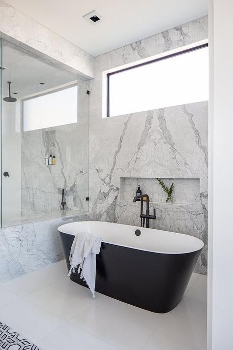 Elegant bathroom boasts a modern black and white oval bathtub with a matte black floor mount tub filler positioned on a white tiled floor beneath a marble tiled niche framed by a bookmatched marble slab wall finished with a picture window. Window Above Bathtub, Tiled Bathtub, Tiled Niche, Tub Bathroom Ideas, Shampoo Niche, Concrete Bathtub, Transitional Bathroom Design, Country Style Bathrooms, Oval Bathtub