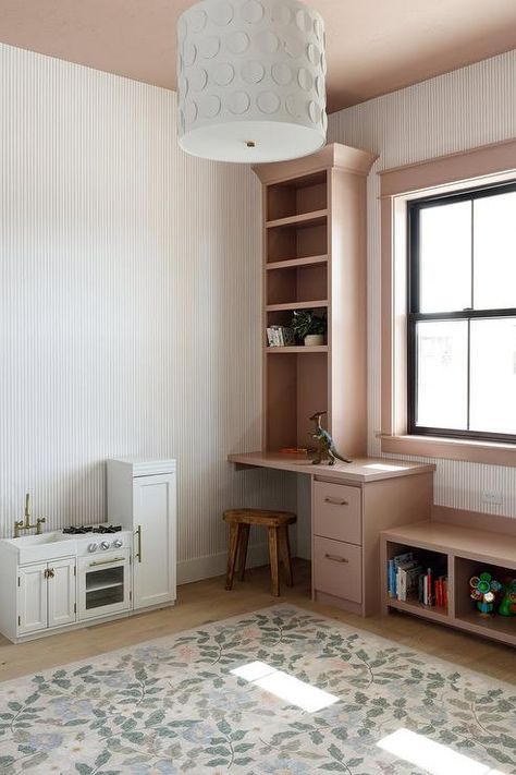 Kid's room features a pink built in window seat next to a built in desk under shelves and a white kitchenette with a floral rug lit by a white pendant hung from a pink ceiling. White Kitchenette, White Family Rooms, Flat Front Cabinets, Built In Desk And Shelves, Pink Girls Room, Blue Stripe Wallpaper, Built In Window Seat, Pink Ceiling, Neutral Kids Room