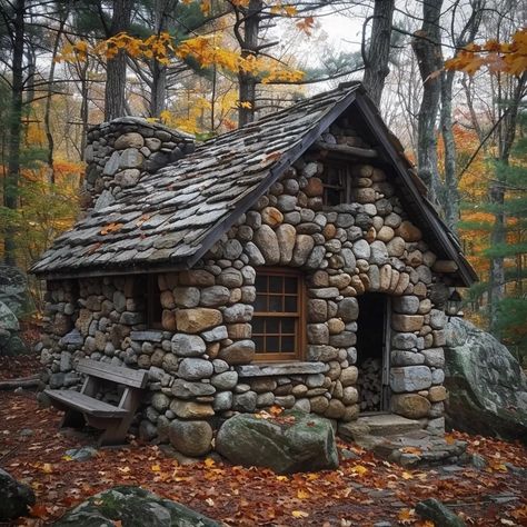 The image is of a small, rustic stone cottage nestled in a forest. The cottage has a fieldstone foundation and walls with a chimney made of the same material. The roof is made of rough-cut wooden shakes. The front door is made of wood. There is a small window on the right side of the cottage. The cottage is surrounded by trees and shrubs. The leaves on the trees are turning brown and orange, which suggests that it is autumn. Cozy Stone Cottage, Slate Roof Cottage, Stone House In Forest, Stone And Log Homes, Small Stone Cottage Interior, Stone Tiny House, Tiny Log Cabin Plans, Small Cottage Exterior Ideas, Stone Foundation House