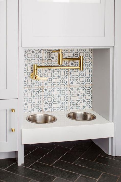 A brass swing arm pot filler is mounted to white and blue mosaic backsplash tiles over a built-in dog food bowl fixed above black herringbone floor tiles and between light gray-blue cabinets with glass and brass pulls. Black Herringbone Floor, Gray Quartz Countertops, Dog Station, Dog Food Station, Transitional Laundry Room, Mudroom Flooring, Grey Floor Tiles, Hydration Station, Bathroom Cabinetry