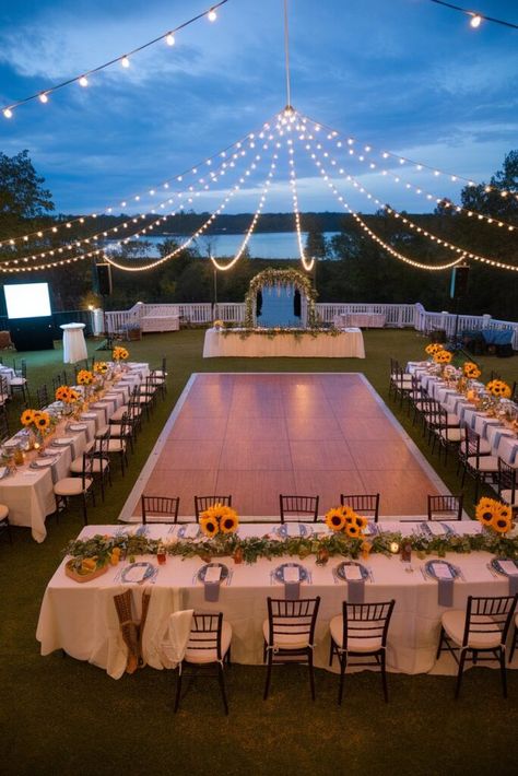 An outdoor wedding reception setup with a dance floor in the center, surrounded by tables decorated with sunflowers and greenery. String lights hang above, creating a romantic atmosphere. A scenic view of water and trees is in the background. Small Modern Wedding Ideas, Outdoor Diy Wedding Reception, Wedding Ideas Minimal, Small Wedding Reception Ideas Outdoor, Garden Party Small Wedding, Small Outdoor Wedding Ideas Backyards, Good Wedding Ideas, Medium Sized Wedding Ideas, Wedding Ideas Location