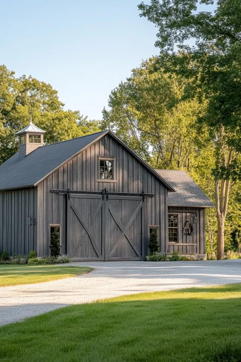 Restored barn in grey board and batten siding natural wood accents large front barn door side prches concrete driveway grass landscape trees in. Unleash your inner farmer and learn how barn houses blend rustic charm with modern living, making them the cozy, unique hideaways you never knew you wanted. Board And Batten Siding Barndominium, Rustic Siding Exterior, Barn Colors, Barn Shop Ideas, Outside Barn Doors Sheds, Simple Horse Barns, Gray Pole Barn With Black Doors, Barnwood Builders Houses, Rustic Barn Homes