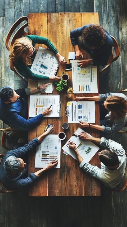 Four professionals are gathered around a wooden table, deeply engaged in a strategy meeting. Various documents, charts, and digital devices are spread out before them, showing they are analyzing data and discussing business strategies. Each participant, displaying focus and collaboration, contributes to the conversation, suggesting the discussion is pivotal. The overhead view captures the teamwork and complexity of their task, emphasizing their commitment to achieving a mutual goal. Team Leader Aesthetic, Business Meeting Aesthetic, Teamwork Aesthetic, Team Work Pictures, Teamwork Photography, Business Man Photography, Business Shooting, Leading A Team, Professional Meeting