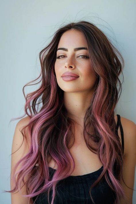 Woman with long, wavy hair dyed in shades of brown and pink, wearing a black top against a plain background. Pink Hair On Brunette Dark Brown, Brown And Rose Gold Balayage, Pink Lowlights In Black Hair, Dark Hair Color With Money Piece, Dark Brown Hair With Rose Gold Money Piece, Dusty Pink Ombre Hair, Hint Of Pink Hair, Black Hair With Rose Gold Money Piece, Rose Gold Highlights Brunette Dark Brown Balayage Hair