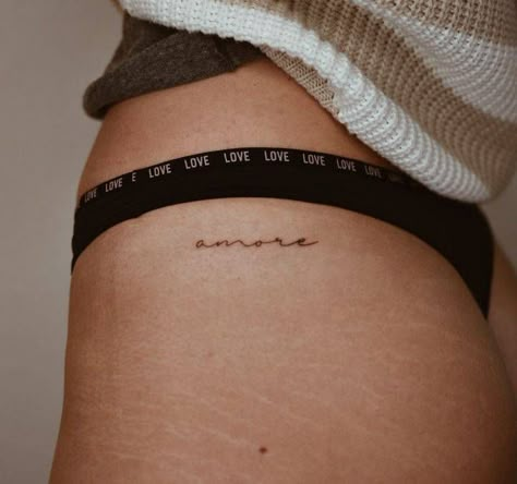 Tattoo of the word "amore" located on the hip. Amor Tattoo On Hip, Amor Hip Tattoo, Danty Tattoos Small On Hip, Hip Writing Tattoos Women, Hip Area Tattoo, One Word Hip Tattoo, Hidden Hip Tattoo, Thats Amore Tattoo, Words Hip Tattoo