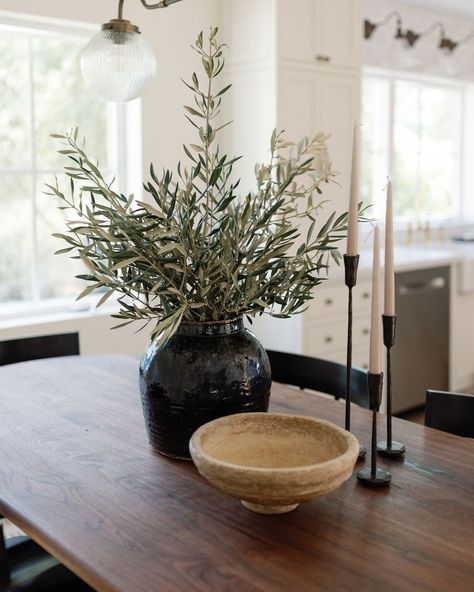 Image Details: black glazed textured vase with olive stems, wood bowl, and black iron taper candle holders on a dark brown wood table. Black Dining Decor, Dark Academia Dining Table Decor, Dining Room Black Table Decor, Candle Holders Dining Table, Greenery On Black Tablecloth, Dining Table Decor Dark Wood, Dark Wood Kitchen Table Decor, Dark Vases Decor, Edgy Farmhouse Decor