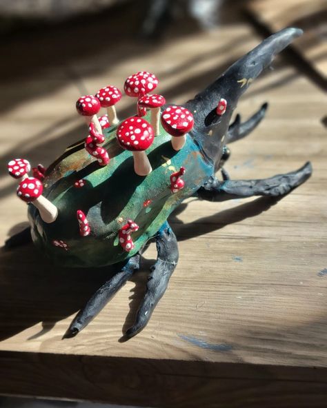 Large beetle with black head, black legs, and green body, facing away from camera, topped with red and white spotted mushrooms. Small gold flecks are scattered across the beetles body, some in the shapes of fae folk wings Paper Mache Beetle, Polymer Clay Beetle, Ceramic Beetle, Dirt Magic, Clay Beetle, Clay Insects, Clay Bugs, Strawberry Animals, Hercules Beetle