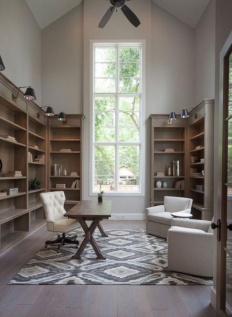 Amazing home office features a tall vaulted ceiling adorned with a ceiling fan over brown stained tall bookcases illuminated by black and gold swing-arm sconces. High Ceiling Office, Transitional Home Office, Home Build Ideas, Office Built Ins, Meteor Garden 2018, Library Office, Office Design Ideas, Office Layout, Office Library