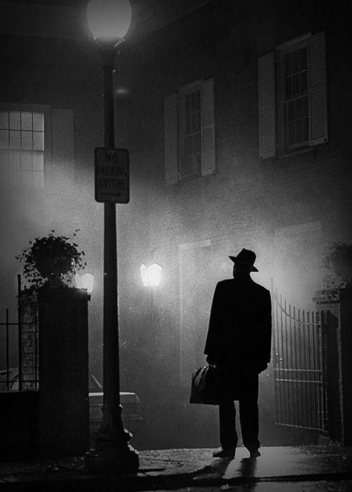 a man in a suit and hat is standing on the street at night with his back turned to the camera