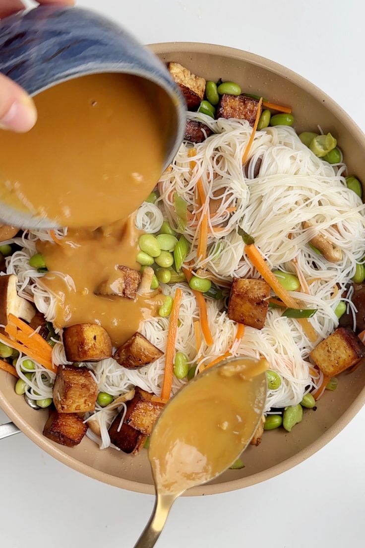 a person pouring sauce into a bowl filled with noodles, peas and croutons