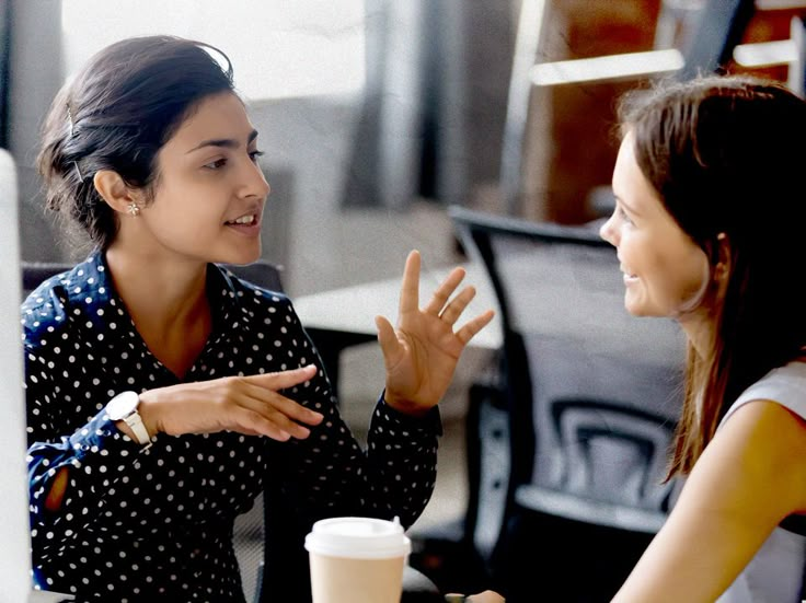 two women sitting at a table talking to each other in front of a computer screen