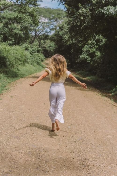 a woman walking down a dirt road with her arms outstretched in the air and trees behind her