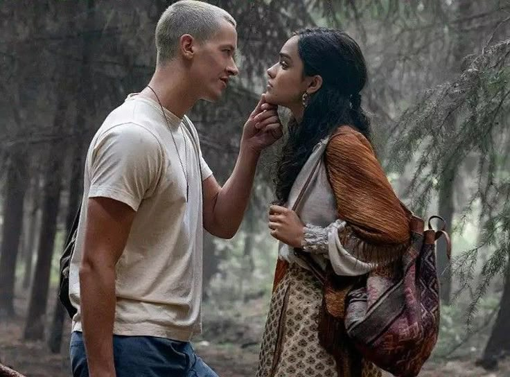 a man and woman standing in the woods looking at each other's eyes as they talk