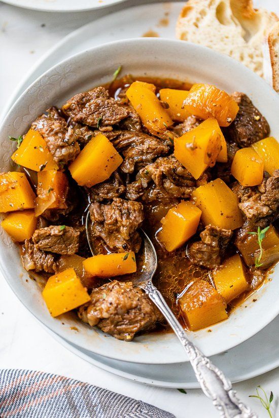 a white bowl filled with meat and squash next to slices of bread on a table