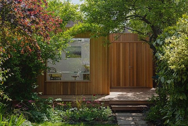 a small wooden house surrounded by trees and flowers in the sunlit garden with steps leading up to it