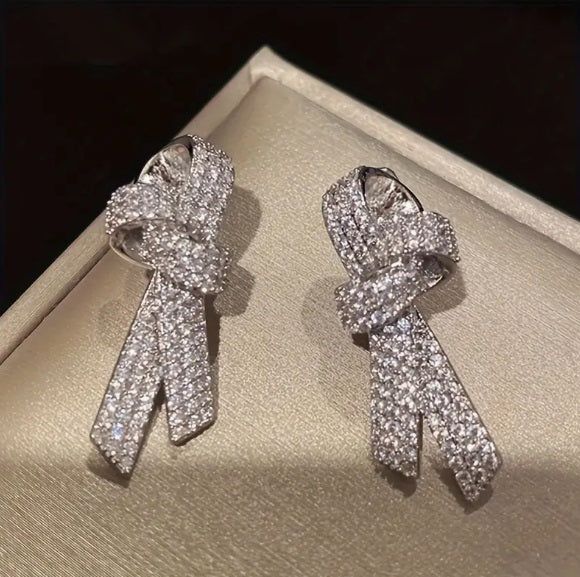 This pair of delicate ribbon bow earrings in a shimmering silver tone, adorned with sparkling gemstones that capture the essence of April's birthstone color. These earrings are crafted with meticulous attention to detail, offering a touch of elegance and sophistication to any ensemble. Each earring features a charming ribbon bow design, exuding femininity and grace. Crafted with nickel-free plating, these earrings are gentle on sensitive skin, ensuring comfort and peace of mind throughout the da Silver Bow Earrings For Anniversary, Silver Earrings With Decorative Bow For Wedding, Elegant Silver Bow Earrings, Glamorous Silver Earrings With Bow, Silver Wedding Earrings With Decorative Bow, Glamorous Silver Bow Earrings, Silver Earrings With Decorative Bow For Evening, Silver Evening Earrings With Decorative Bow, Elegant White Gold Earrings With Bow