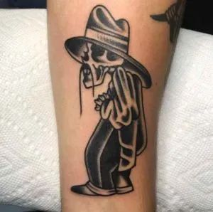 a skeleton wearing a hat and holding a pipe tattoo on the leg, with a bird flying above it