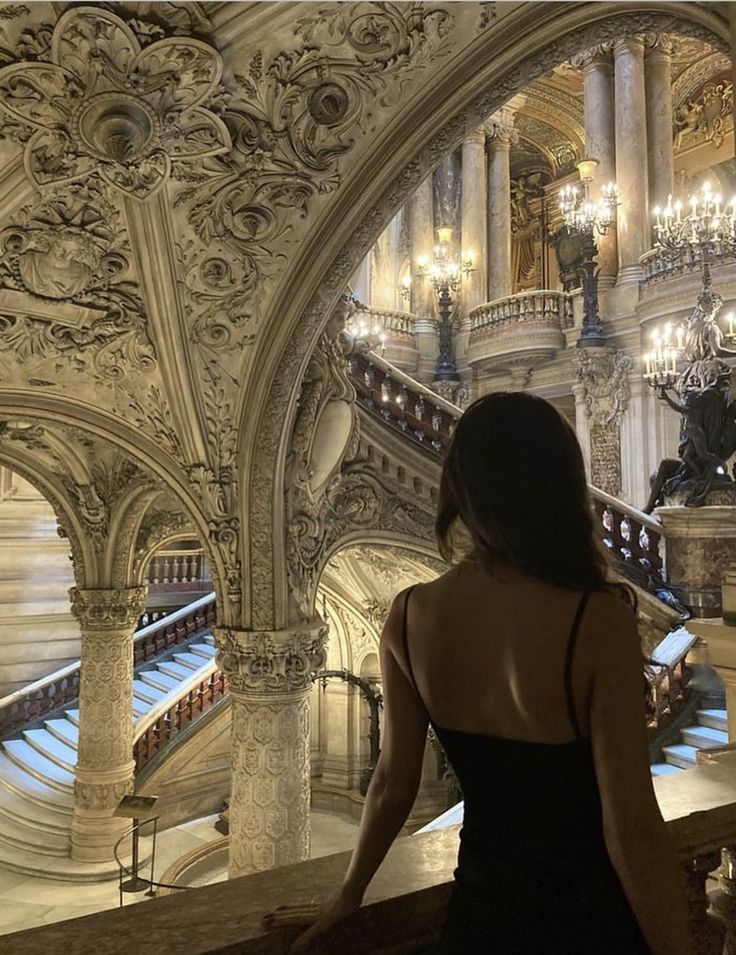 a woman in a black dress is looking down at the stairs and chandeliers