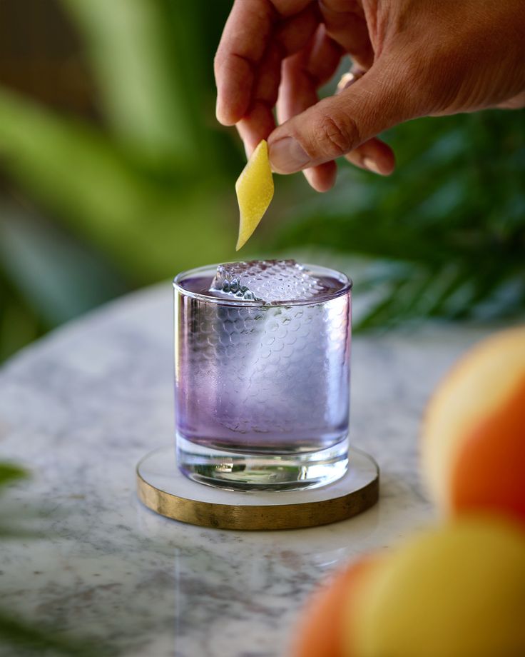 a person holding a lemon wedge over a glass filled with purple liquid on top of a marble table