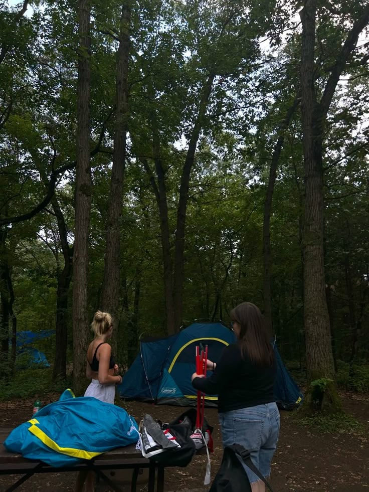 two women sitting at a picnic table with a tent in the background and trees behind them