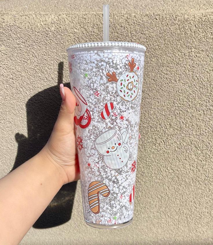 a hand is holding a cup with frosting and christmas decorations on it, next to a wall