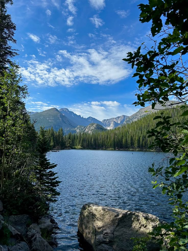 a lake surrounded by trees and mountains under a blue sky with clouds in the background