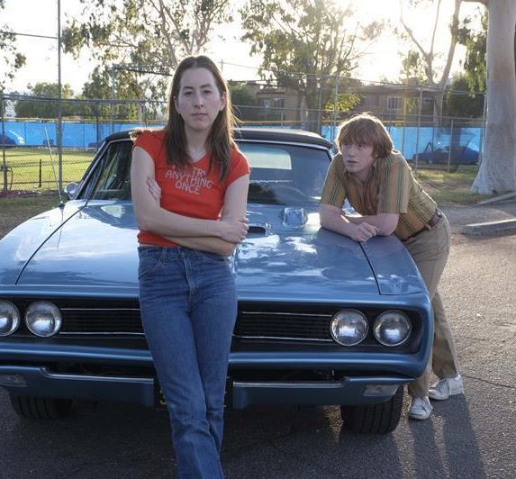 two people sitting on the hood of a car with their arms crossed and one person standing next to it