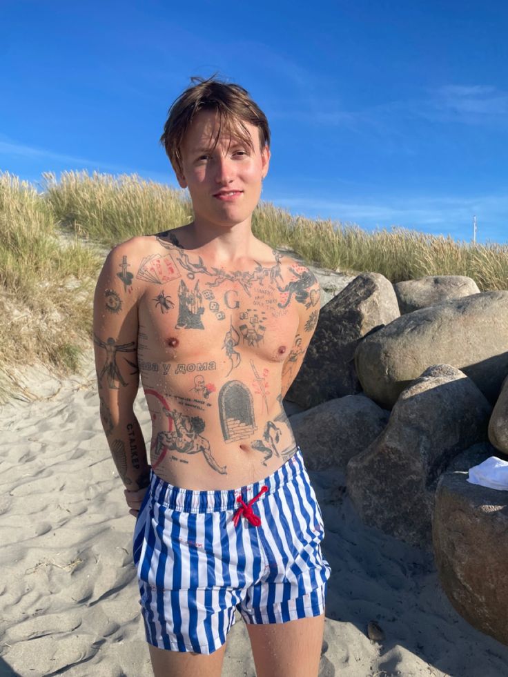 a man with tattoos on his chest standing in front of some rocks and sand at the beach