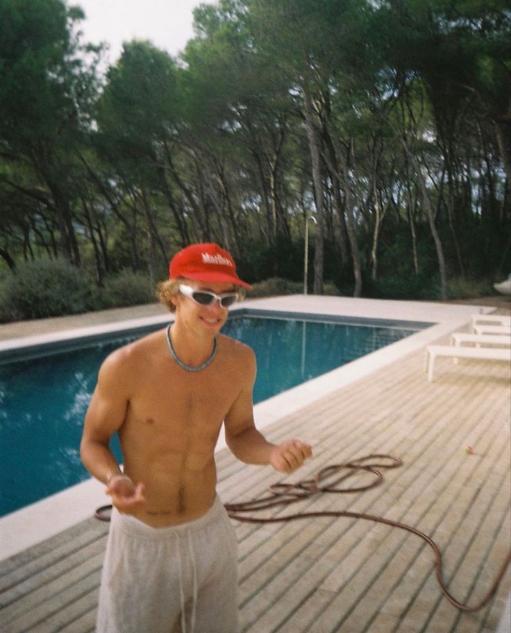 a man with no shirt on standing next to a swimming pool holding a water hose