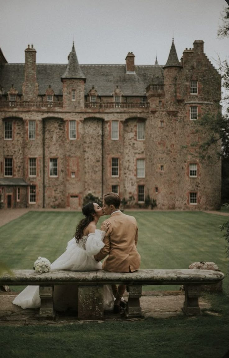 a bride and groom sitting on a bench in front of an old castle like building