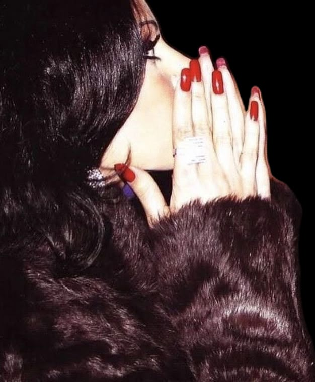 a woman with red nails is holding her hand up to her face while wearing a fur coat