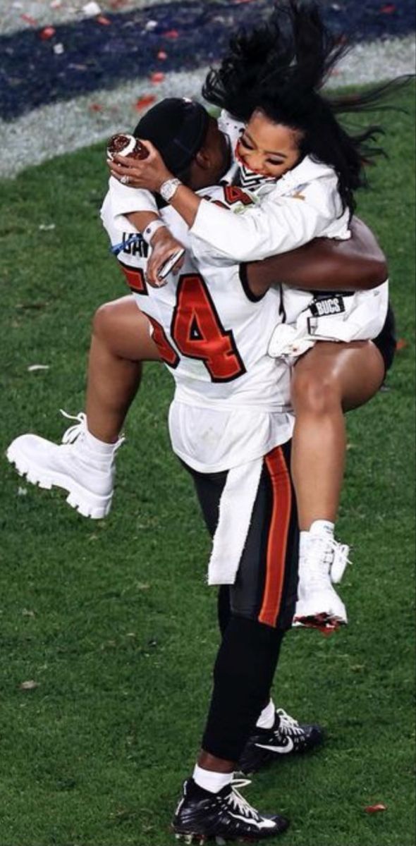 two women are hugging on the field during a football game, one is wearing an orange and white uniform