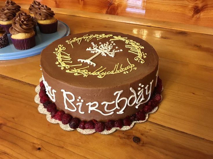 a chocolate birthday cake sitting on top of a wooden table next to cupcakes