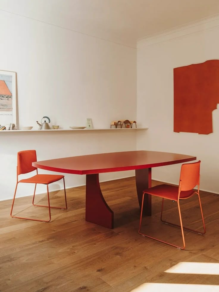 a red table and two orange chairs in a white room with wood flooring on the side