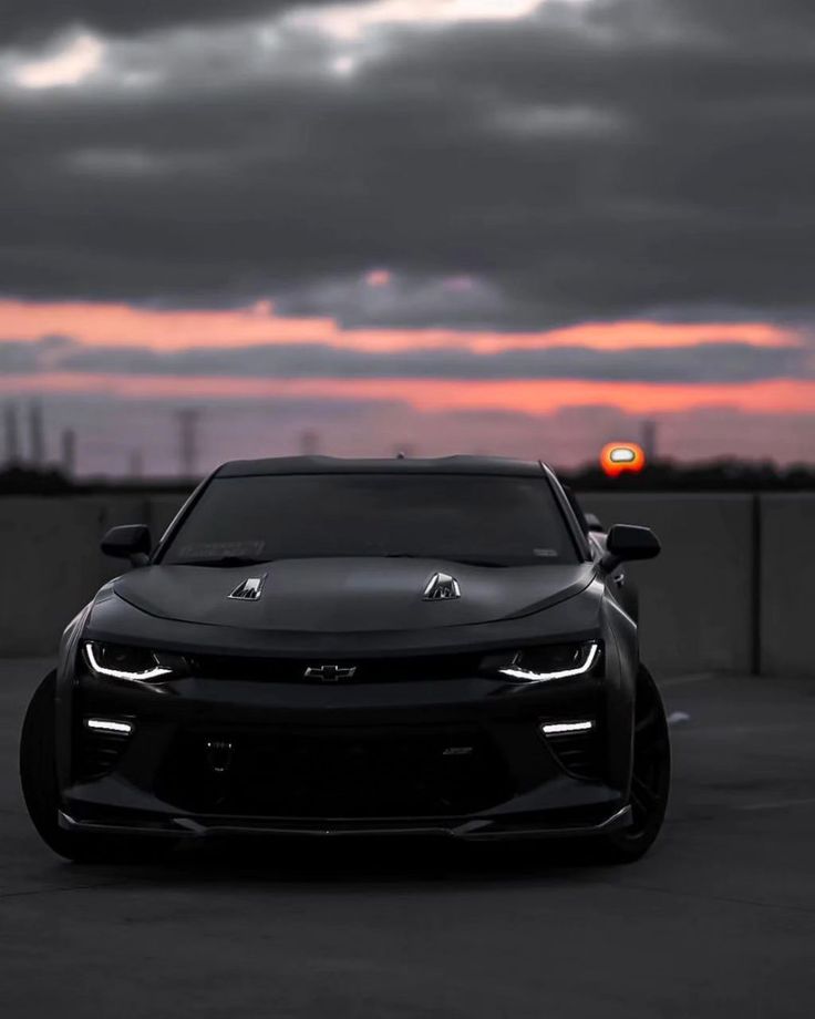 the front end of a black sports car parked in a parking lot at dusk with dark clouds